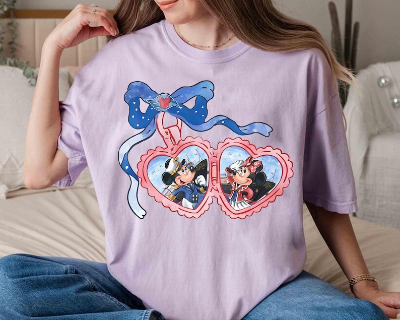 May include: A lavender t-shirt with a graphic of Mickey Mouse and Minnie Mouse in a heart-shaped locket. The locket is decorated with a blue bow and a heart.