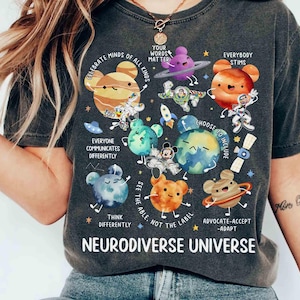 Op de afbeelding: Een donkergrijs T-shirt met een kleurrijk grafisch ontwerp met planeten, sterren en de tekst "Celebrate Minds of All Kinds", "Your Words Matter", "Everybody Stims", "Everyone Communicates Differently", "Choose to Include", "See the Able, Not the Label", "Think Differently", "Advocate-Accept-Adapt" en "Neurodiverse Universe".