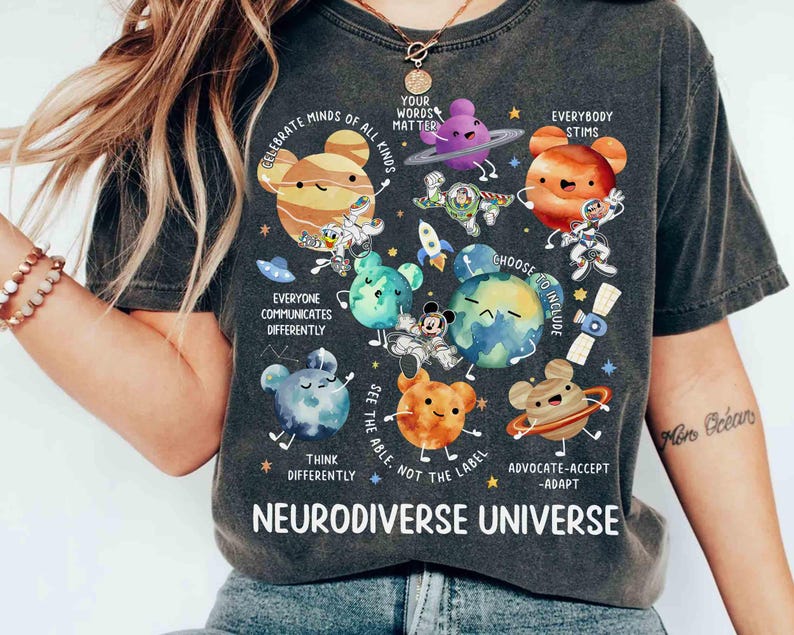 Op de afbeelding: Een donkergrijs T-shirt met een kleurrijk grafisch ontwerp met planeten, sterren en de tekst "Celebrate Minds of All Kinds", "Your Words Matter", "Everybody Stims", "Everyone Communicates Differently", "Choose to Include", "See the Able, Not the Label", "Think Differently", "Advocate-Accept-Adapt" en "Neurodiverse Universe".