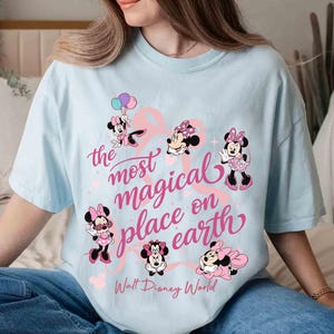 K&ouml;nnte beinhalten: Hellblaues T-Shirt mit mehreren Minnie-Mouse-Illustrationen in Rosa und Schwarz. Der Text "the most magical place on earth" ist in rosa Schrift geschrieben, darunter "Walt Disney World".