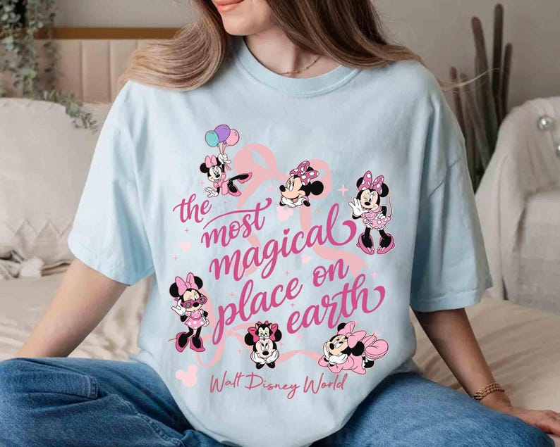 K&ouml;nnte beinhalten: Hellblaues T-Shirt mit mehreren Minnie-Mouse-Illustrationen in Rosa und Schwarz. Der Text "the most magical place on earth" ist in rosa Schrift geschrieben, darunter "Walt Disney World".