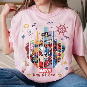 May include: A light pink t-shirt featuring a colorful graphic of Marvel characters and nautical elements. The design includes the text "Marvel Day At Sea" and various cartoon characters. The shirt has short sleeves.