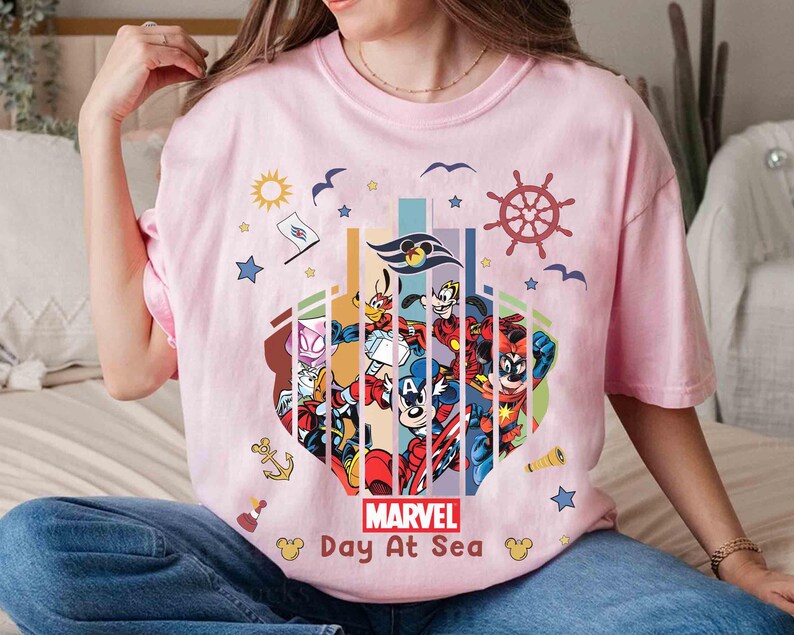 May include: A light pink t-shirt featuring a colorful graphic of Marvel characters and nautical elements. The design includes the text "Marvel Day At Sea" and various cartoon characters. The shirt has short sleeves.