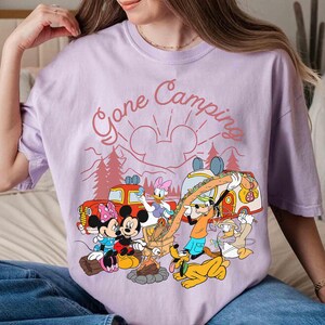 Pu&ograve; includere: Maglietta lilla chiaro con una grafica di campeggio con Topolino, Minnie, Paperino, Paperina, Pippo e Pluto. Il testo "Gone Camping" &egrave; sopra i personaggi. Il design include un fal&ograve;, alberi e un camper.