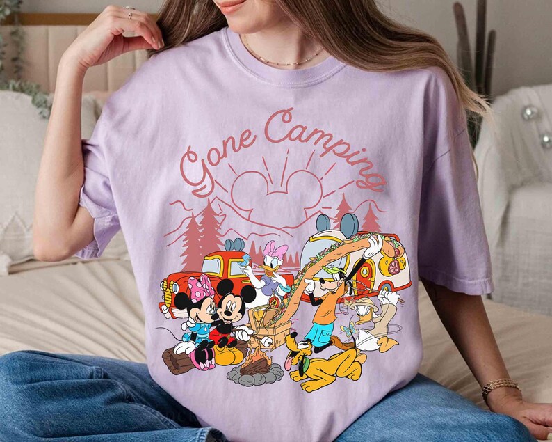 Pu&ograve; includere: Maglietta lilla chiaro con una grafica di campeggio con Topolino, Minnie, Paperino, Paperina, Pippo e Pluto. Il testo "Gone Camping" &egrave; sopra i personaggi. Il design include un fal&ograve;, alberi e un camper.