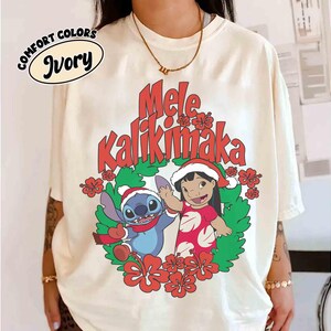 May include: Ivory-colored Comfort Colors t-shirt featuring a festive design. The graphic includes the text "Mele Kalikimaka" in red, along with cartoon characters Stitch and Lilo in Santa hats, surrounded by a wreath of flowers.