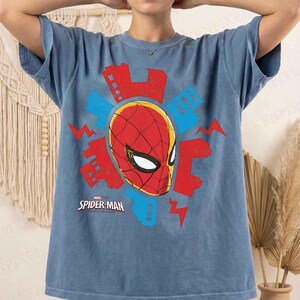 Puede incluir: Una camiseta azul de manga corta con un gr&aacute;fico de la m&aacute;scara de Spider-Man en rojo y negro. El dise&ntilde;o incluye siluetas de edificios urbanos estilizados en rojo y azul y formas de rayos, con el texto Spider-Man Across the Spider-Verse.