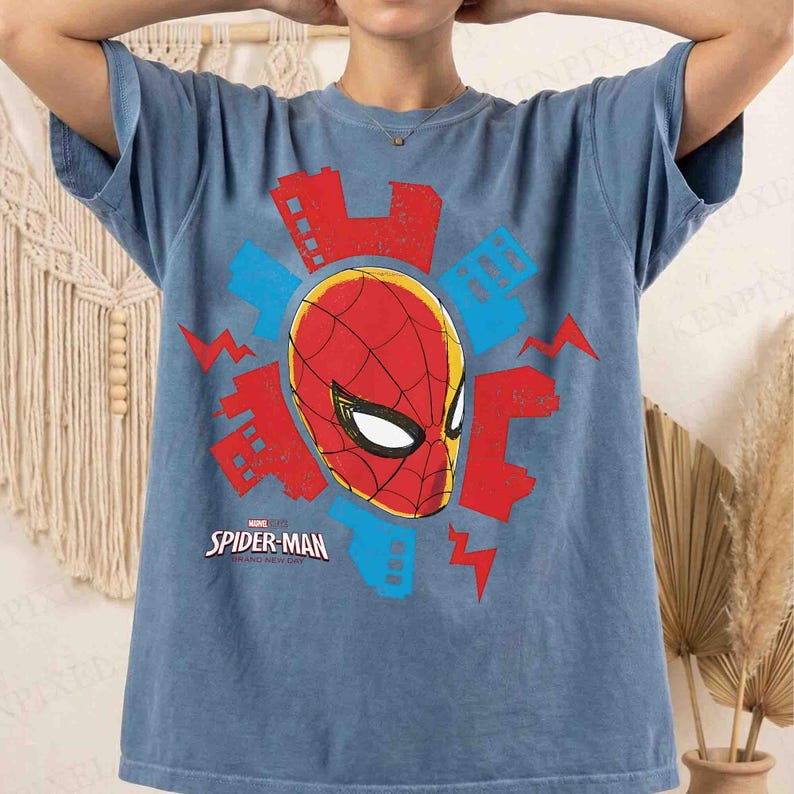 Puede incluir: Una camiseta azul de manga corta con un gr&aacute;fico de la m&aacute;scara de Spider-Man en rojo y negro. El dise&ntilde;o incluye siluetas de edificios urbanos estilizados en rojo y azul y formas de rayos, con el texto Spider-Man Across the Spider-Verse.
