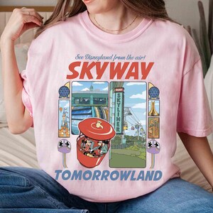 Puede incluir: Camiseta rosa claro con un dise&ntilde;o gr&aacute;fico que presenta las palabras "SKYWAY" y "TOMORROWLAND". El dise&ntilde;o incluye una g&oacute;ndola roja con Mickey y Minnie Mouse, e im&aacute;genes de las atracciones de Disneyland. La camiseta tiene cuello redondo y mangas cortas.