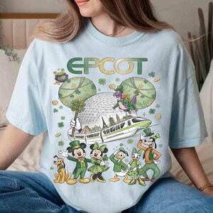 May include: Pale blue t-shirt with a green "EPCOT" logo and a St. Patrick's Day themed Disney graphic. The design includes Mickey and Minnie Mouse, Donald Duck, Goofy, and a dragon, with a monorail and the EPCOT sphere.