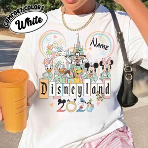 May include: White t-shirt featuring a Disneyland 2020 design with Mickey and Minnie Mouse, Donald and Daisy Duck, Goofy, and Pluto. The shirt has the text "Disneyland" in a colorful font, with the words "Comfort Colors" and "White" in a circle.