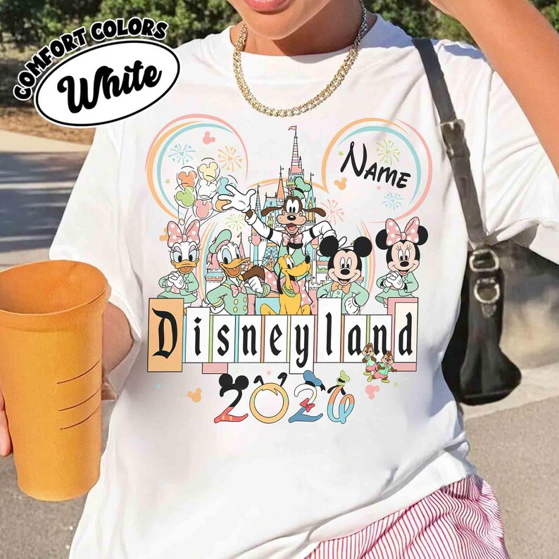 May include: White t-shirt featuring a Disneyland 2020 design with Mickey and Minnie Mouse, Donald and Daisy Duck, Goofy, and Pluto. The shirt has the text "Disneyland" in a colorful font, with the words "Comfort Colors" and "White" in a circle.