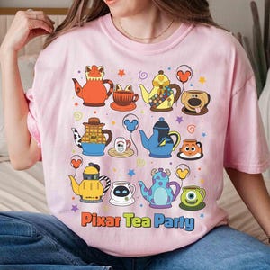 May include: Pink t-shirt featuring a colorful graphic of cartoon teapots and teacups. The design includes characters from various animated films, with the text "Pixar Tea Party" at the bottom. The shirt is made of a soft material.