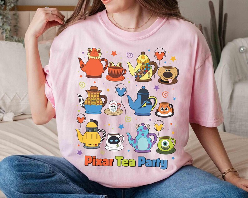 May include: Pink t-shirt featuring a colorful graphic of cartoon teapots and teacups. The design includes characters from various animated films, with the text "Pixar Tea Party" at the bottom. The shirt is made of a soft material.