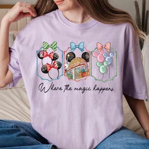May include: A light purple t-shirt featuring three framed illustrations. The illustrations include Minnie Mouse ears, a theme park ride, and balloons. The text "Where the magic happens" is written below the illustrations.