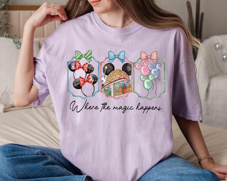 May include: A light purple t-shirt featuring three framed illustrations. The illustrations include Minnie Mouse ears, a theme park ride, and balloons. The text "Where the magic happens" is written below the illustrations.