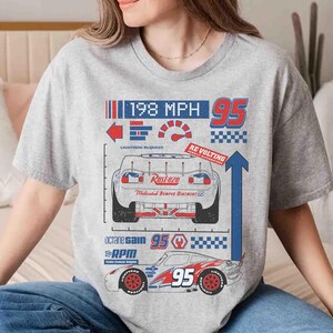 K&ouml;nnte beinhalten: Ein graues, meliertes T-Shirt mit einem Grafikdruck eines Rennwagens in R&uuml;ck- und Seitenansicht. Das Design enth&auml;lt den Text 198 MPH, 95, Rust-eze, Lightning McQueen, Re-volting, Octane Gain und RPM sowie verschiedene Rennsport-Symbole.