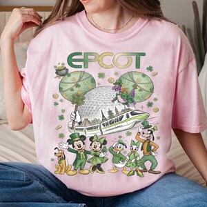 May include: Pale pink t-shirt with a St. Patrick's Day design featuring the word "EPCOT". The graphic includes Disney characters Mickey and Minnie Mouse, Donald and Daisy Duck, and Goofy in green outfits, plus a monorail.