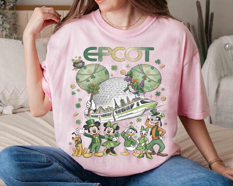 May include: Pale pink t-shirt with a St. Patrick's Day design featuring the word "EPCOT". The graphic includes Disney characters Mickey and Minnie Mouse, Donald and Daisy Duck, and Goofy in green outfits, plus a monorail.