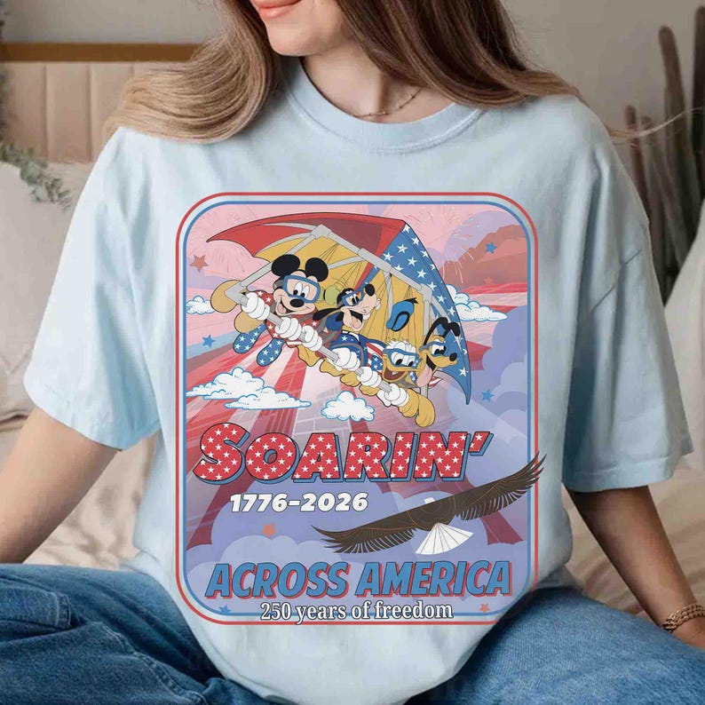 May include: Light blue t-shirt featuring Mickey Mouse, Goofy, and Donald Duck soaring on a hang glider with an American flag design. The shirt has the words "SOARIN'" and "ACROSS AMERICA" with the years 1776-2026 and "250 years of freedom". An eagle is also featured.