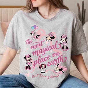 K&ouml;nnte beinhalten: Ein graues T-Shirt mit mehreren Minnie-Maus-Illustrationen in Rosa und Schwarz. Der Text "the most magical place on earth" ist in rosa Schrift geschrieben, darunter "Walt Disney World".