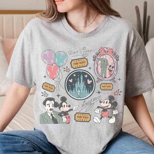 Peut inclure: T-shirt gris avec un motif graphique color&eacute; repr&eacute;sentant Mickey Mouse, un ch&acirc;teau, des ballons et les textes "Walt Disney" et "Mickey Mouse". Le motif comprend des illustrations de billets de parc et un portrait de Walt Disney avec Mickey.