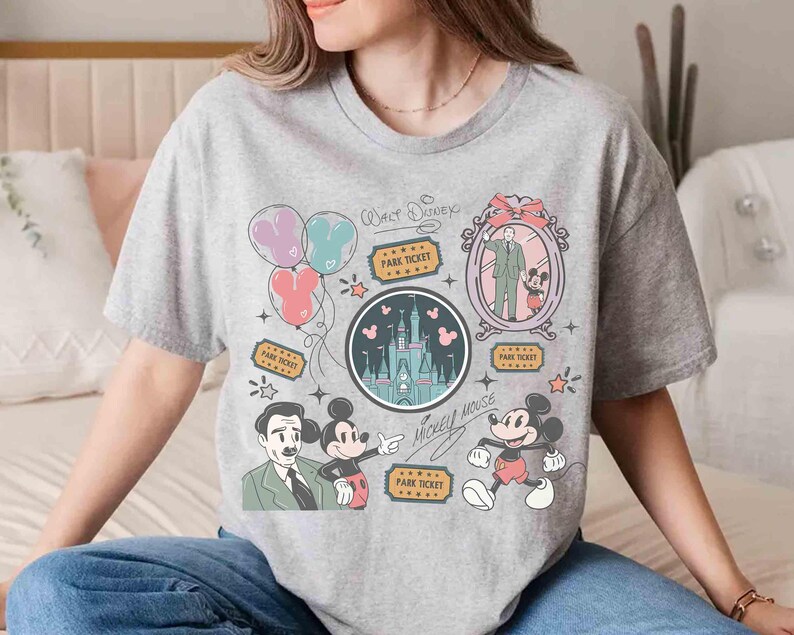 Peut inclure: T-shirt gris avec un motif graphique color&eacute; repr&eacute;sentant Mickey Mouse, un ch&acirc;teau, des ballons et les textes "Walt Disney" et "Mickey Mouse". Le motif comprend des illustrations de billets de parc et un portrait de Walt Disney avec Mickey.
