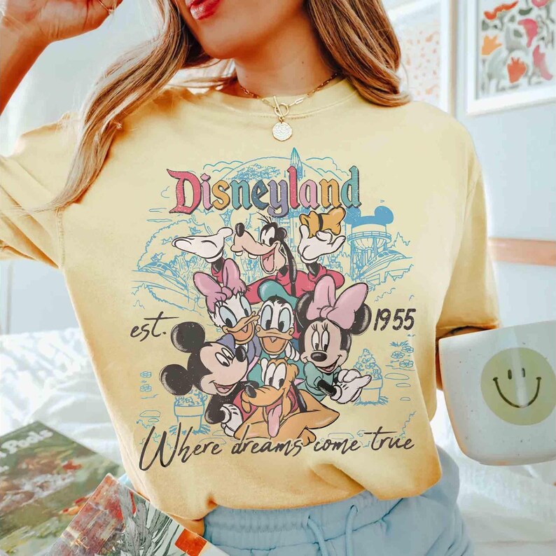 May include: Yellow t-shirt featuring the word "Disneyland" in a retro font, along with illustrations of Mickey Mouse, Minnie Mouse, Donald Duck, Daisy Duck, Goofy, and Pluto. The shirt also includes the text "est. 1955" and "Where dreams come true."