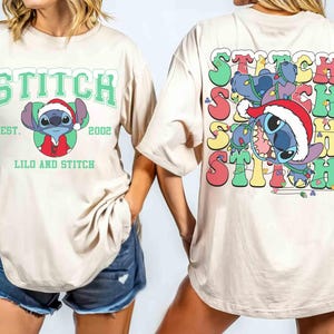 May include: Beige t-shirt featuring a graphic of Stitch wearing a Santa hat, with the text "STITCH" above and "LILO AND STITCH" below. The back of the shirt has a colorful Stitch design with the word "STITCH" repeated.