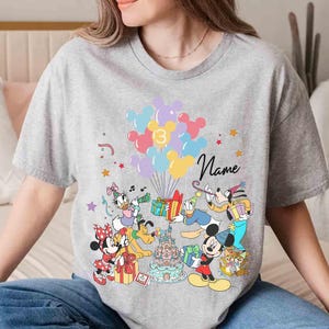 May include: A gray t-shirt featuring a colorful graphic of Disney characters, balloons, and a birthday cake. The number "3" is on the balloons, and the word "Name" is written in cursive. Mickey Mouse, Minnie Mouse, and Donald Duck are among the characters.