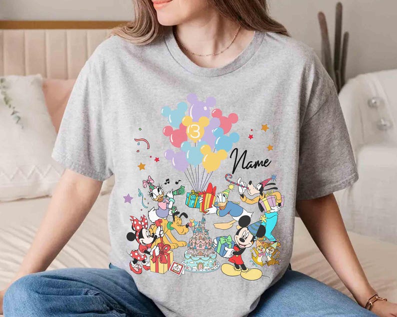 May include: A gray t-shirt featuring a colorful graphic of Disney characters, balloons, and a birthday cake. The number "3" is on the balloons, and the word "Name" is written in cursive. Mickey Mouse, Minnie Mouse, and Donald Duck are among the characters.