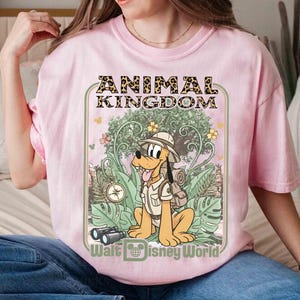 Puede incluir: Camiseta rosa claro con un dise&ntilde;o de Pluto con ropa de safari, con el texto "ANIMAL KINGDOM" y "Walt Disney World". El dise&ntilde;o incluye una br&uacute;jula, binoculares y un fondo de selva.