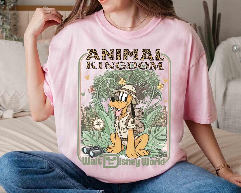 Puede incluir: Camiseta rosa claro con un dise&ntilde;o de Pluto con ropa de safari, con el texto "ANIMAL KINGDOM" y "Walt Disney World". El dise&ntilde;o incluye una br&uacute;jula, binoculares y un fondo de selva.