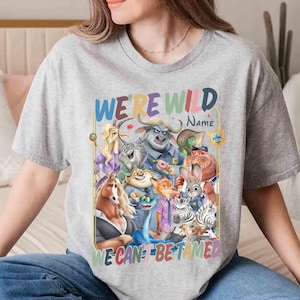 May include: A gray t-shirt featuring a colorful graphic of various cartoon animal characters. The text "WE'RE WILD" is arched above the image, with "NAME" below it. The phrase "WE CAN'T BE TAMED" is at the bottom.