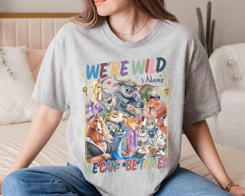 May include: A gray t-shirt featuring a colorful graphic of various cartoon animal characters. The text "WE'RE WILD" is arched above the image, with "NAME" below it. The phrase "WE CAN'T BE TAMED" is at the bottom.