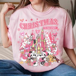 May include: Pink t-shirt with a Christmas graphic featuring Disney characters and the text "CHRISTMAS ON MAIN STREET". The design includes Mickey and Minnie Mouse, Donald Duck, Goofy, and a castle.