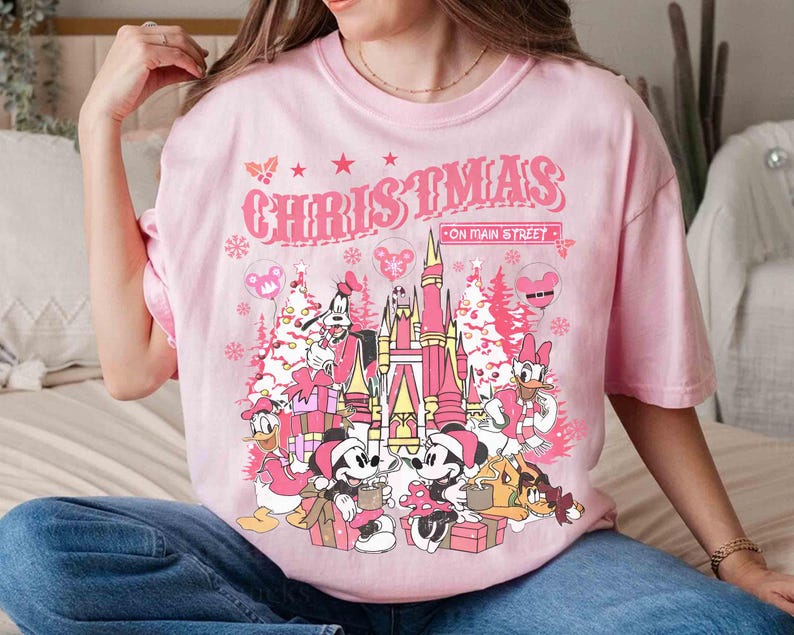 May include: Pink t-shirt with a Christmas graphic featuring Disney characters and the text "CHRISTMAS ON MAIN STREET". The design includes Mickey and Minnie Mouse, Donald Duck, Goofy, and a castle.