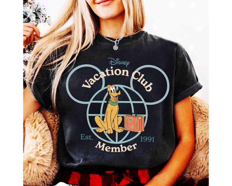 Puede incluir: Camiseta negra con el logo de Disney Vacation Club, Pluto, una maleta y el texto "Member Est. 1991." El dise&ntilde;o incluye orejas de Mickey Mouse en azul claro y un globo terr&aacute;queo. La camiseta tiene las mangas dobladas.