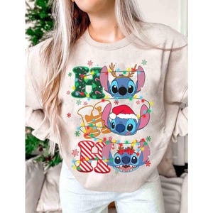 May include: Beige sweatshirt with a colourful graphic featuring the words "HO HO HO" and three cartoon characters. The characters are decorated with Christmas lights and festive accessories, including antlers and a Santa hat.