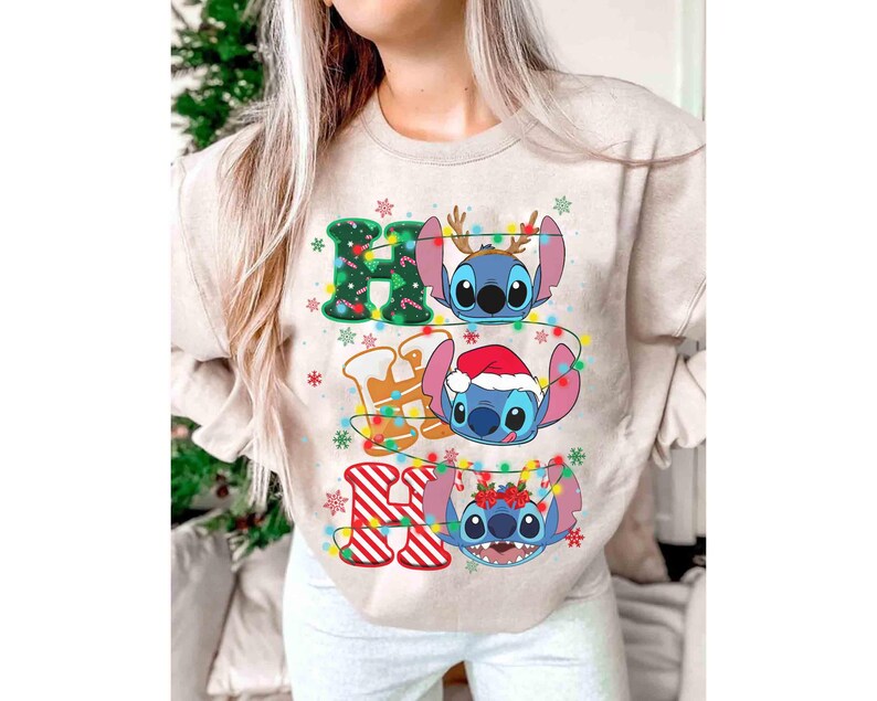 May include: Beige sweatshirt with a colourful graphic featuring the words "HO HO HO" and three cartoon characters. The characters are decorated with Christmas lights and festive accessories, including antlers and a Santa hat.