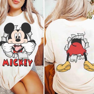 May include: Cream-colored t-shirt featuring a graphic of Mickey Mouse bursting through a torn wall, with the word "Mickey" in red. The back of the shirt shows Mickey's legs and shoes. A fun, cartoon-themed apparel item.