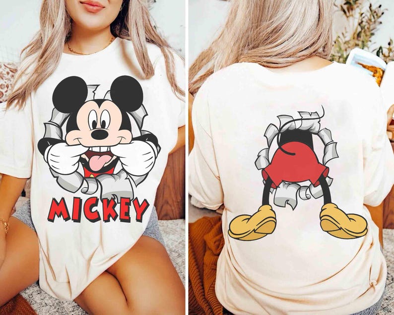 May include: Cream-colored t-shirt featuring a graphic of Mickey Mouse bursting through a torn wall, with the word "Mickey" in red. The back of the shirt shows Mickey's legs and shoes. A fun, cartoon-themed apparel item.