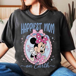 Pode incluir: Camiseta cinza escuro com Minnie Mouse, com la&ccedil;o rosa e vestido de bolinhas. O texto "Happiest Mom on Earth" est&aacute; em azul. O desenho est&aacute; dentro de um oval rosa e azul.