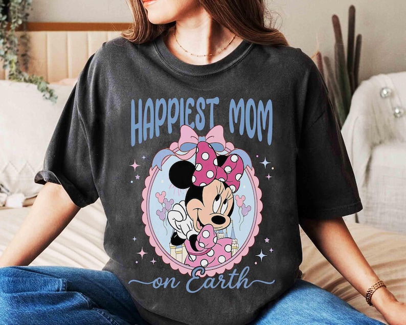 Pode incluir: Camiseta cinza escuro com Minnie Mouse, com la&ccedil;o rosa e vestido de bolinhas. O texto "Happiest Mom on Earth" est&aacute; em azul. O desenho est&aacute; dentro de um oval rosa e azul.