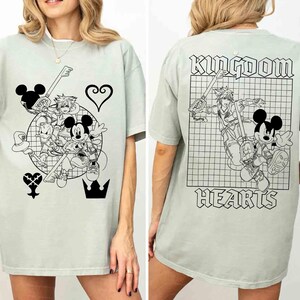 Puede incluir: Camiseta verde claro con un gr&aacute;fico en blanco y negro de Mickey Mouse y otros personajes con armas en forma de llave. La parte trasera de la camiseta tiene las palabras "Kingdom Hearts".