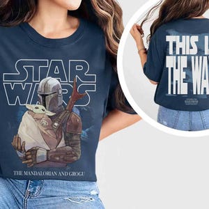 May include: A navy blue t-shirt featuring a Star Wars graphic on the front with The Mandalorian holding Grogu and the text The Mandalorian and Grogu. The back of the shirt displays the white text This Is The Way with a Star Wars logo below it.
