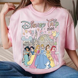 May include: A light pink t-shirt featuring the text "Disney Trip 2026" with a graphic of Disney princesses and iconic park imagery. The design includes Cinderella, Ariel, Belle, and Jasmine. The shirt has a crew neck and short sleeves.