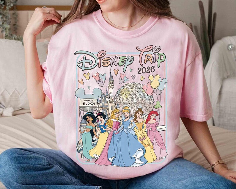 May include: A light pink t-shirt featuring the text "Disney Trip 2026" with a graphic of Disney princesses and iconic park imagery. The design includes Cinderella, Ariel, Belle, and Jasmine. The shirt has a crew neck and short sleeves.