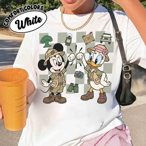 May include: White t-shirt with a graphic of Minnie Mouse and Daisy Duck in safari outfits, fist-bumping. The design includes a checkered background with camping and travel-themed icons. The text "COMFORT COLORS White" is printed at the top.