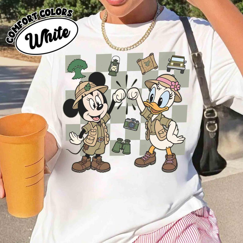 May include: White t-shirt with a graphic of Minnie Mouse and Daisy Duck in safari outfits, fist-bumping. The design includes a checkered background with camping and travel-themed icons. The text "COMFORT COLORS White" is printed at the top.